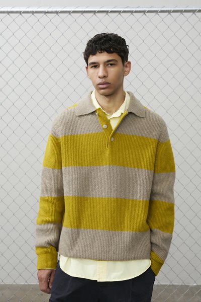 Mladi moški s kodrastimi temnimi lasmi, oblečen v rumen in bež črtast pleten pulover čez svetlo rumeno srajco, stoji ob ograji iz kovinske mreže.
