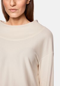 Helle beige Bluse mit langen Ärmeln, weitem Ausschnitt und weicher, glatter Textur. Verfügt über überschnittene Schultern und minimale Nähte.
