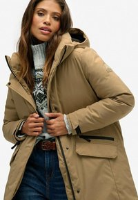 Beige geïsoleerde parka met een capuchon, zwarte accenten, zijzakken en een ritssluiting, gedragen over een gemêleerd grijs truitje en blauwe spijkerbroek.