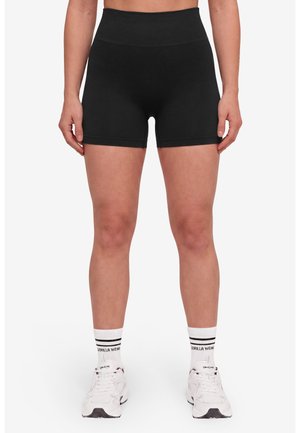 Vrouw die zwarte, hooggetailleerde, aansluitende shorts draagt, witte sportsokken met zwarte strepen en witte sneakers, staand tegen een effen achtergrond.
