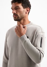 Hellgrauer Pullover mit gerippten Bündchen, rundem Halsausschnitt und einer Reißverschlusstasche am linken Ärmel. Glatte Stoffstruktur.