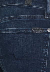 Jeans de mezclilla de color azul oscuro con una superficie texturizada, que presentan un patrón de costura en zigzag en el bolsillo trasero y una pequeña etiqueta de la marca.