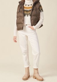 Hnědá péřová vesta přes krémový svetr s vzorovanou yoke, bílé kalhoty s manžetami a béžové kotníkové boty se zipem.