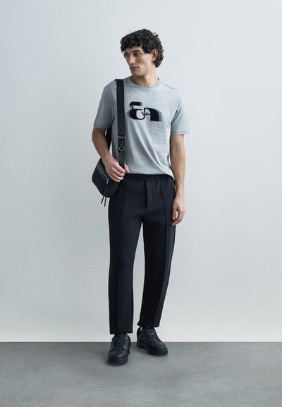 Sivý tričko s čiernym grafickým dizajnom, v kombinácii s čiernymi elegantnými teplákmi. Doplnky zahŕňajú čiernu kabelku cez rameno a čierne tenisky.