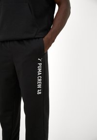 Man in zwarte trainingsbroek met het logo "PUMA CREW '48" op de linkerdij tegen een witte achtergrond, met zijn hand in zijn broekzak.