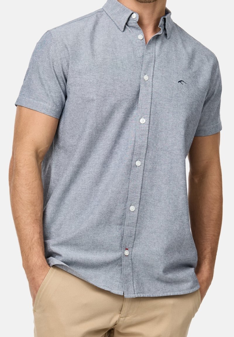 Homme portant une chemise gris clair à manches courtes avec un petit logo noir sur la poitrine et un pantalon beige, les mains dans les poches.