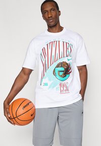 Λευκό βαμβακερό T-shirt με πολύχρωμο γραφικό των Memphis Grizzlies, που απεικονίζει το λογότυπο μιας αρκούδας. Κρατούνται γκρι σορτς Nike και πορτοκαλί μπάλα μπάσκετ.