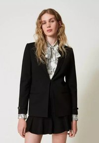 Jonge vrouw met golvend blond haar, gekleed in een zwarte blazer, een patroon blouse in het wit met strik, en een zwarte geplisseerde rok, tegen een effen achtergrond.