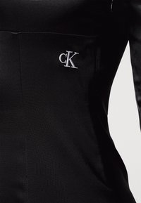 Černé šaty s hladkým materiálem a obtáhnutým střihem. Vyšívané logo "CK" na levé straně. Minimalistický styl s čistými liniemi.