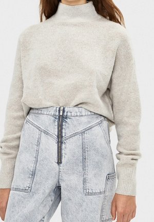 Kobieta ubrana w beżowy sweter z golfem wpuszczony w wysokiej talii, jasnoniebieskie dżinsy z przednim zamkiem błyskawicznym i dużymi kieszeniami.
