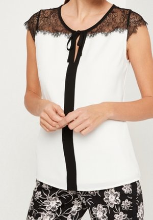 Blouse blanche sans manches avec empiècement en dentelle noire, rayure verticale noire et nœud au col, associée à un pantalon à motif floral noir et blanc.