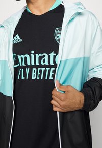 Czarna sportowa koszulka z turkusowymi akcentami, z napisem "Emirates Fly Better". Lekka kurtka na wierzchu, z turkusowym i czarnym wzorem w bloki kolorów.