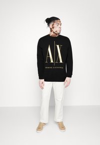 Sort sweatshirt med guld "AX" logo og teksten "ARMANI EXCHANGE", parret med hvide bukser og beige sneakers, mod en ensfarvet baggrund.