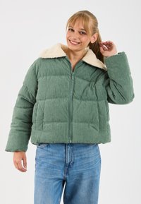 Smiling woman adjusting her ponytail wearing a green corduroy puffer jacket with a cream fleece collar and blue jeans.