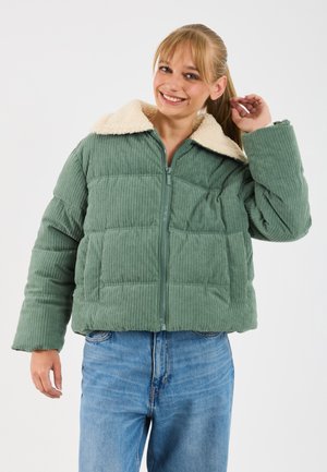 Glunderende vrouw die haar paardenstaart aanpast, gekleed in een groene corduroy pufferjack met een crèmekleurige fleece kraag en blauwe jeans.