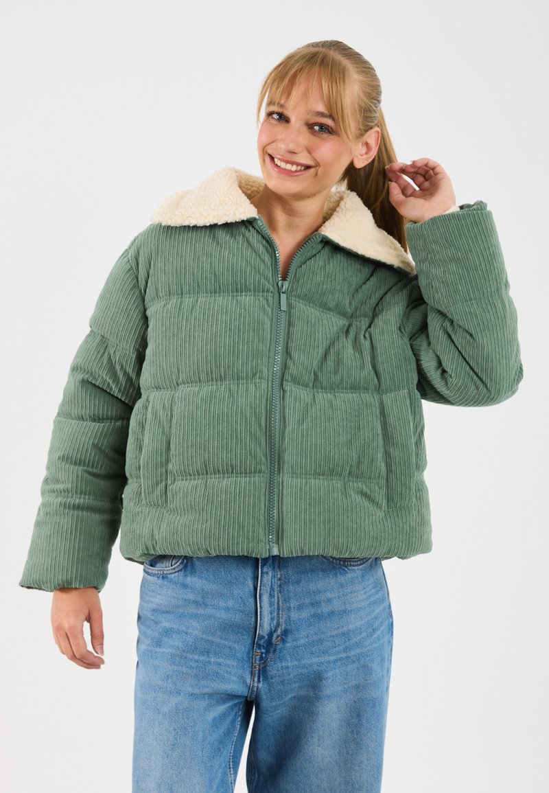 Smiling woman adjusting her ponytail wearing a green corduroy puffer jacket with a cream fleece collar and blue jeans.
