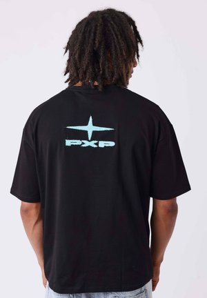 Persoon met dreadlocks die een zwart t-shirt draagt met een lichtblauwe ster en de tekst "PXP" op de rug, staand tegen een effen achtergrond.