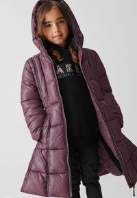 Lång lila puffjacka med huva, med kviltade sektioner, framdragkedja och sidofickor. Bärs över en svart hoodie och byxor.