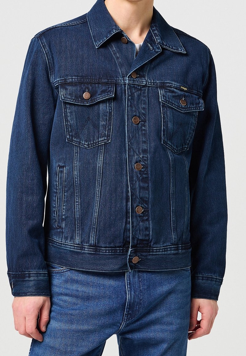 Homme portant une veste en jean bleu foncé avec fermeture à boutons, deux poches poitrine à rabat, assortie à un jean bleu, sur fond uni.