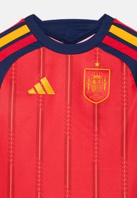 Κόκκινη φανέλα ποδοσφαίρου με μπλε και κίτρινες ρίγες στους ώμους, έμβλημα της εθνικής ομάδας της Ισπανίας με ένα αστέρι, και κίτρινο λογότυπο Adidas στο στήθος.