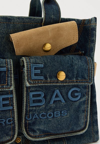 Τσάντα από τζιν με πολλαπλές τσέπες, μπλε κεντημένο κείμενο "The Bag Jacobs" και καφέ φάσα από σουέντ με χρυσά κουμπώματα.