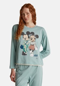 Joven mujer con cabello largo y rojo que lleva una blusa de manga larga azul claro y pantalones, con un gráfico de Mickey y Minnie Mouse acompañado de texto.
