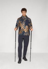 Haut à capuche à manches longues pour homme en motif tie-dye bleu et marron avec une poche avant, associé à un pantalon sombre et des bâtons de randonnée, portant des chaussures noires.