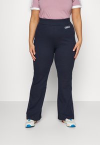 Person som bär svarta Adidas-byxor med logotyp på höften, ihop med vita sneakers och en mauve kortärmad topp mot en enfärgad bakgrund.