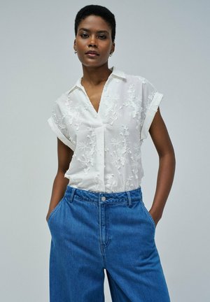Blusa blanca bordada de manga corta con cuello, combinada con pantalones vaqueros azules de talle alto con un corte relajado y bolsillos.