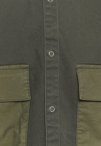 Tummanvihreä paita, jossa on napillinen etuosa, hopeiset yksityiskohdat ja kaksi suurta paikkataskua, jotka on valmistettu kontrastivärisestä oliivinvihreästä kankaasta.