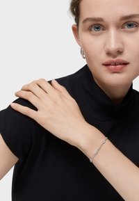 Pulsera de plata con eslabones decorativos, llevada en una muñeca, combinada con una blusa negra de cuello alto. Textura suave y diseño delicado.