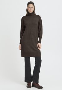 Barna kötött garbóruha hosszú ujjakkal, bordázott szegéllyel és laza szabással, fekete nadrággal és sötétbarna bokacsizmával párosítva.