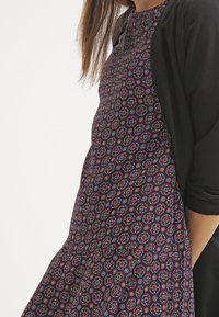 Abito blu navy con un design a pattern caratterizzato da motivi circolari rossi e blu. Accompagnato da un cardigan nero. Tessuto dalla texture liscia.