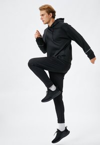 Czarny strój sportowy składający się z bluzy z kapturem i zwężających się spodni dresowych, oba wykonane z gładkiego materiału, z pasującymi czarnymi sneakersami i szarymi akcentami.