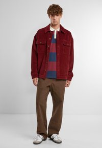 Červená kordurová bunda s prednými vreckami, nosená cez prúžkovanú polo košeľu v červenej, modrej a bielej farbe, spárovaná s hnedými nohavicami a šedými teniskami.