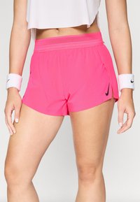 Eine Frau trägt leuchtend pinkfarbene Nike-Sportshorts und weiße Armbänder. Sie steht mit entspannten Händen an den Seiten vor einem schlichten Hintergrund.