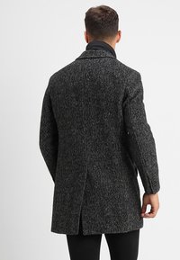 Man in een donkergrijs, gestructureerd wollen jasje met een achteropening, zwarte broek en een hoge kraag zwart kledingstuk daaronder, met zijn rug naar de kijker gekeerd.