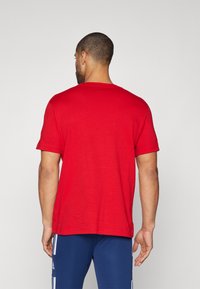 T-shirt rouge à manches courtes avec un col ras du cou, présentant une texture lisse et un ourlet droit. Porté avec un pantalon de sport bleu, visible à la taille.