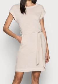 Robe rayée beige clair avec des manches courtes, taille cintrée et poches latérales. Tissu doux et coupe décontractée. Détail de ceinture à la taille.