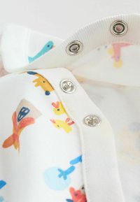 Fehér szövet színes állat- és játékmintákkal, fémes patentokkal a nyakkivágásnál. Lágy textúra és közeli részlet látható.