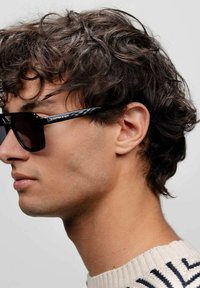 Zwarte zonnebril met een glanzende afwerking, met hoekige monturen en donkere glazen, gedragen door een persoon met krullend haar en een lichte trui.