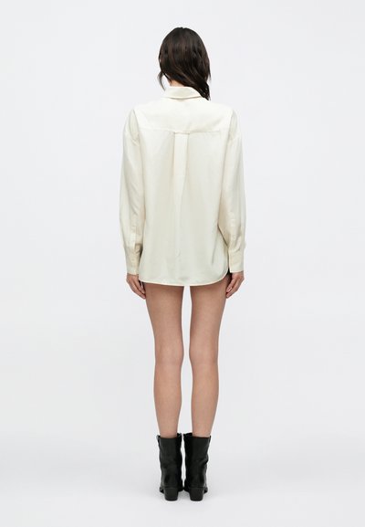 Chemise surdimensionnée de couleur crème avec une texture lisse, manches longues et ourlet arrondi. Portée avec des bottines noires.