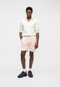 Jeune homme debout, les mains dans les poches, portant une chemise boutonnée vert pâle, un short rose, des chaussettes blanches et des baskets noires.