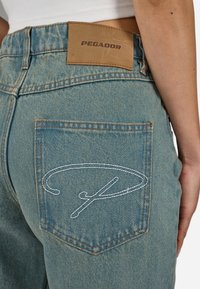 Ljusblå denimjeans har en läderetikett med "PEGADOR" på midjan och dekorativ vit söm på bakfickan.