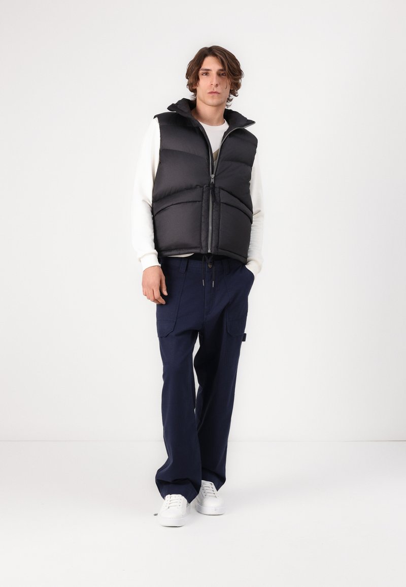 Gilet rembourré noir avec capuche, présentant des détails en matelassage, fermeture éclair, associé à un pantalon cargo large bleu marine et des baskets blanches.