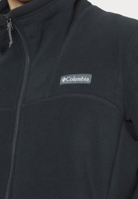 Giacca in pile nera con colletto alto, zip completa e logo Columbia sulla parte superiore sinistra. Texture morbida, design aderente.