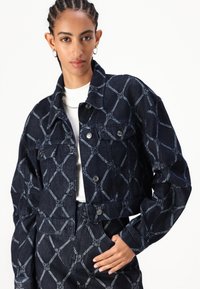 Korte denimjack met een donkere wastint en een geometrisch patroon in blauw en grijs, accenten van zilverkleurige hardware en twee voorkruiken.