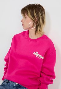 Ružičasti dukserica od mekog materijala s okruglim izrezom, s bijelim "ellesse" logom. Model nosi velike srebrne naušnice.