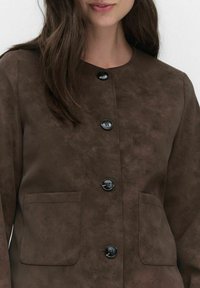 Femme portant une veste en daim brun foncé avec des boutons noirs et deux poches plaquées à l'avant, montrée de la nuque à la taille.