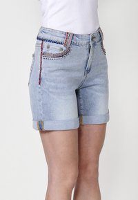 Shorts en denim de couleur bleu clair avec des ourlets retroussés ; décorés de broderies colorées sur les poches et la ceinture ; style classique à cinq poches.